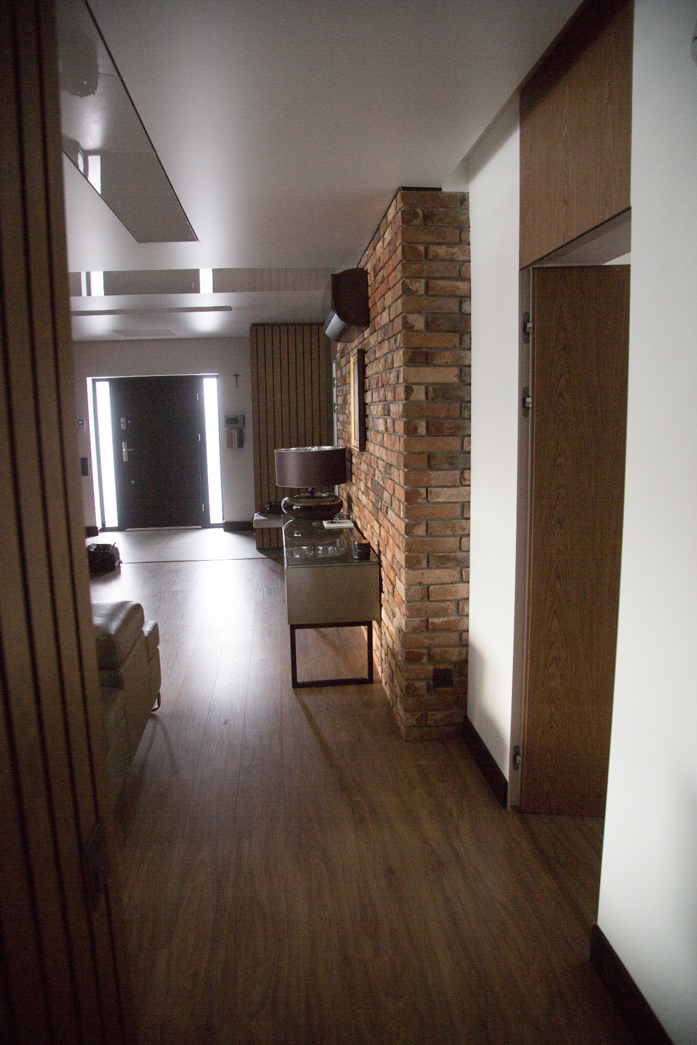 Nowoczesny przedpokój z ceglaną ścianą, drewnianą podłogą i minimalistycznym oświetleniem. Widoczny fragment sofy i drzwi wejściowe z przeszkleniami.