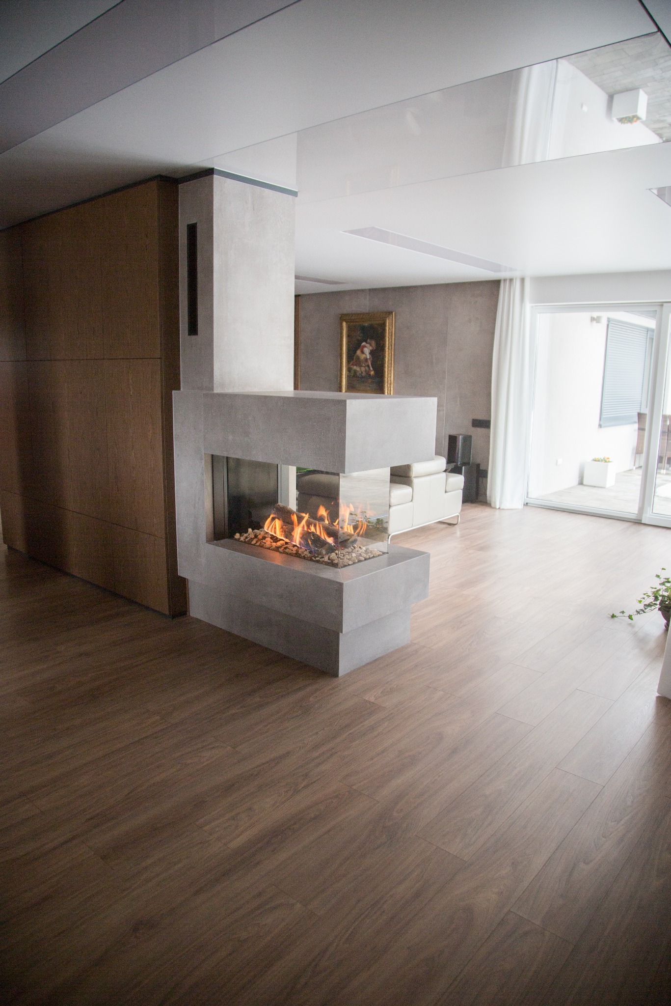 Nowoczesny salon z kominkiem narożnym o minimalistycznej bryle, drewnianą podłogą i jasnymi ścianami. Widoczne detale wykończenia wnętrz i płomienie w kominku.