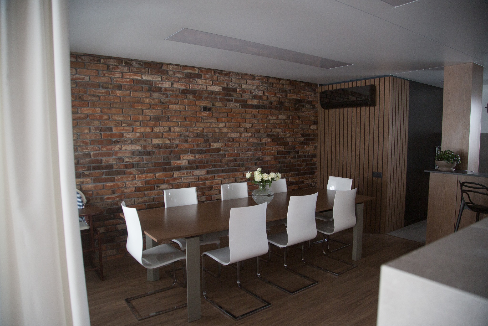 Jadalnia z ceglaną ścianą i nowoczesnymi białymi krzesłami. Na stole wazon z białymi kwiatami. Widoczne drewniane panele ścienne i fragment zasłony.