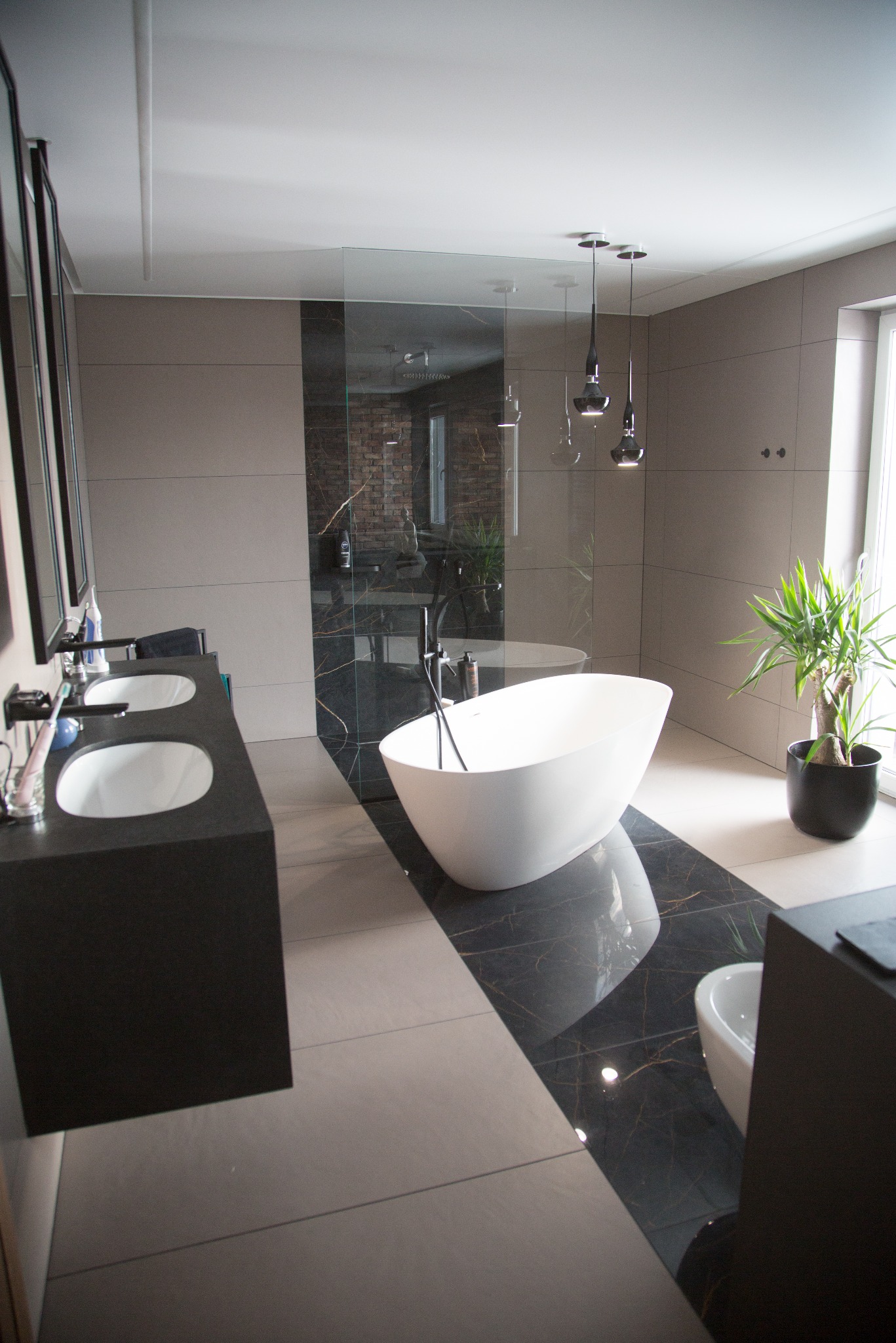 Nowoczesna łazienka z wolnostojącą wanną, czarnym blatem umywalki i marmurową podłogą. Minimalistyczne oświetlenie i roślina doniczkowa dodają elegancji.