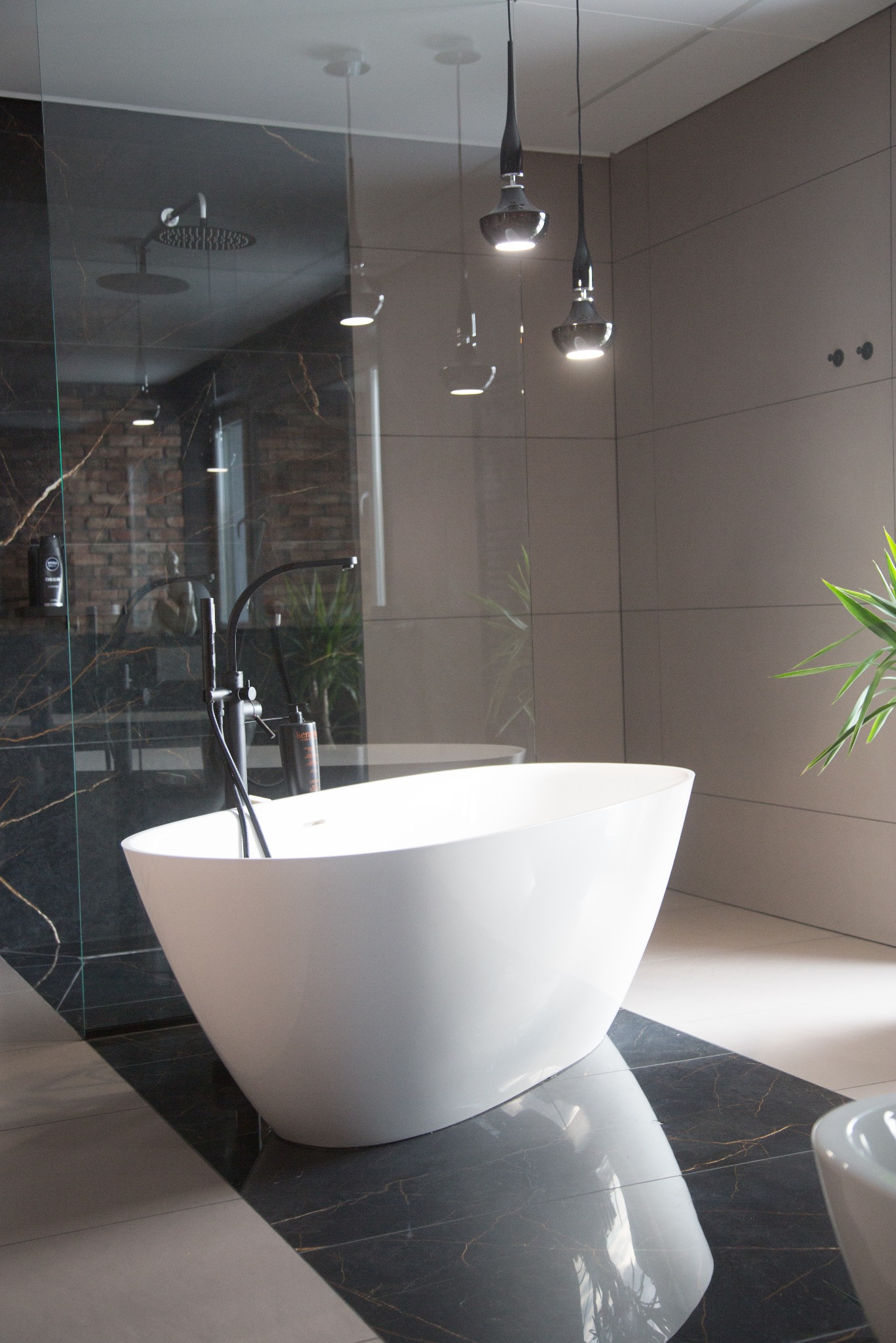 Nowoczesna łazienka z wolnostojącą białą wanną na tle czarnej ściany z marmuru i szklanej kabiny prysznicowej. Minimalistyczne oświetlenie i rośliny doniczkowe.