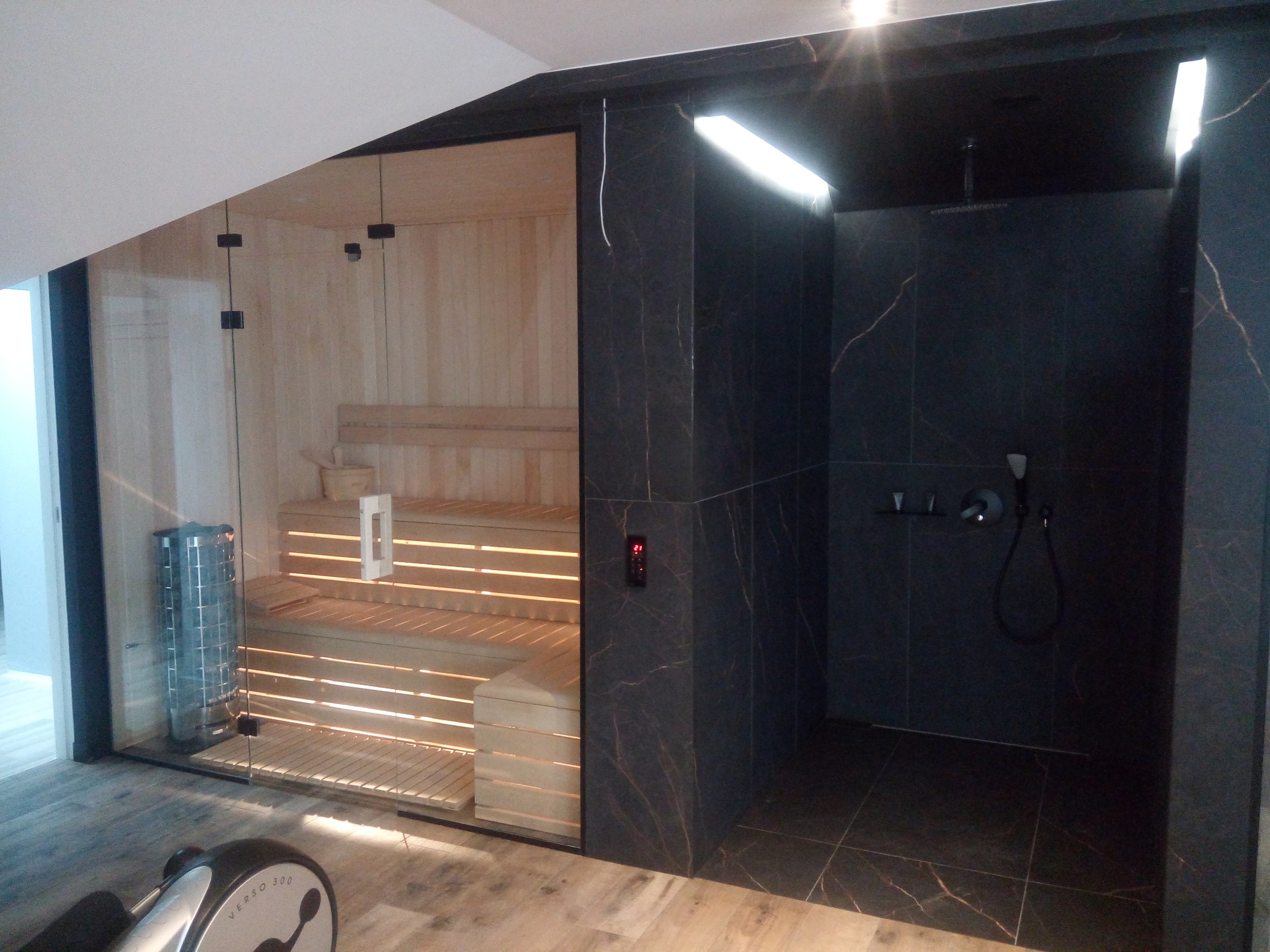 Nowoczesna łazienka z sauną: kabina prysznicowa z ciemnymi płytkami imitującymi marmur i przeszklona sauna z drewnianym wnętrzem, oświetlona ciepłym światłem.