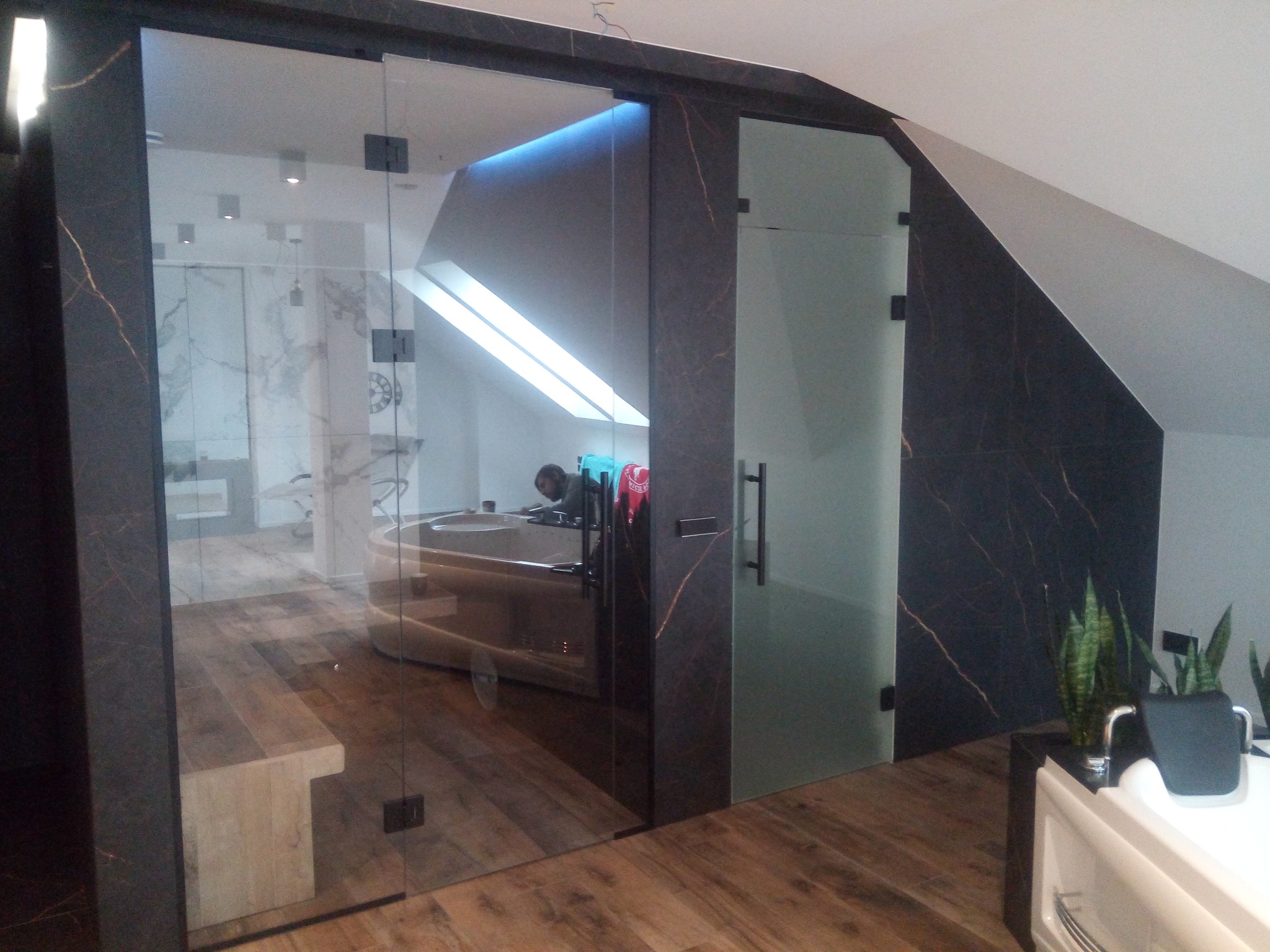 Szklane drzwi przesuwne z czarnymi okuciami, oddzielające łazienkę z wanną od reszty pomieszczenia. Widoczne drewniane podłogi i skośny sufit.