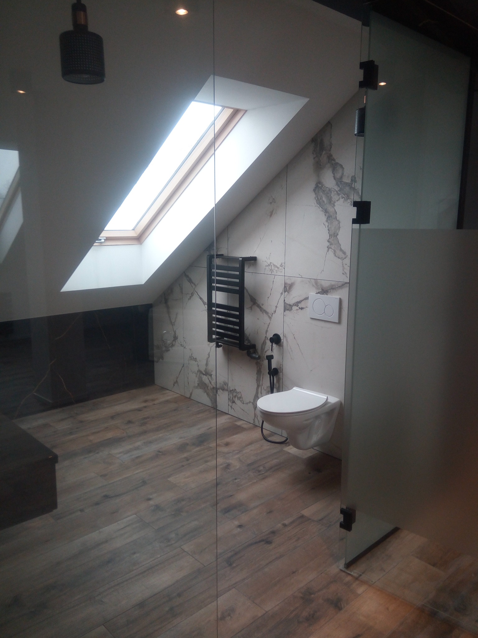 Nowoczesna łazienka na poddaszu z oknem dachowym, marmurowymi płytkami i drewnopodobną podłogą. Widoczna toaleta podwieszana i szklana kabina prysznicowa.