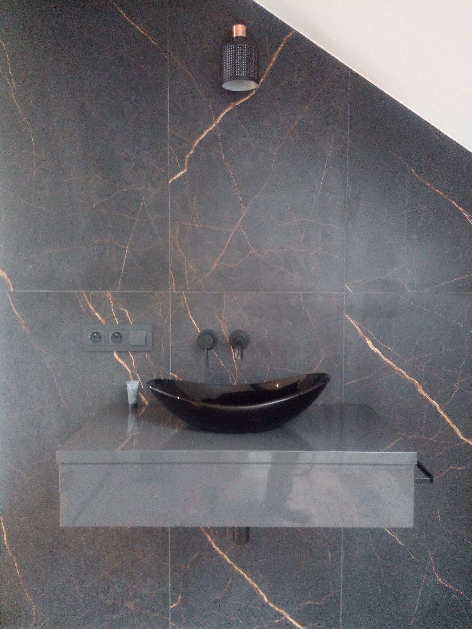 Nowoczesna łazienka z czarną umywalką na tle płytek imitujących marmur z złotymi żyłami. Minimalistyczna armatura i oświetlenie punktowe.