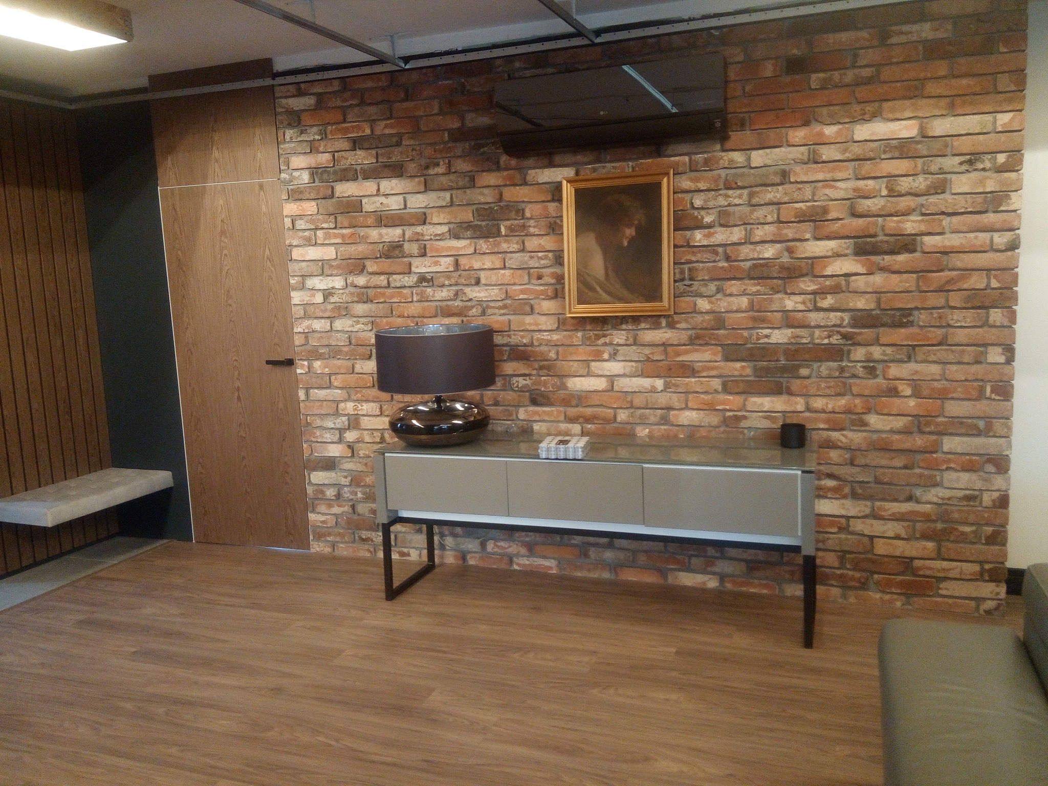 Ceglana ściana z obrazem i szafką RTV, oświetlona lampą. Widoczna drewniana podłoga i fragment sofy. Dekoracyjne wykończenie wnętrza.