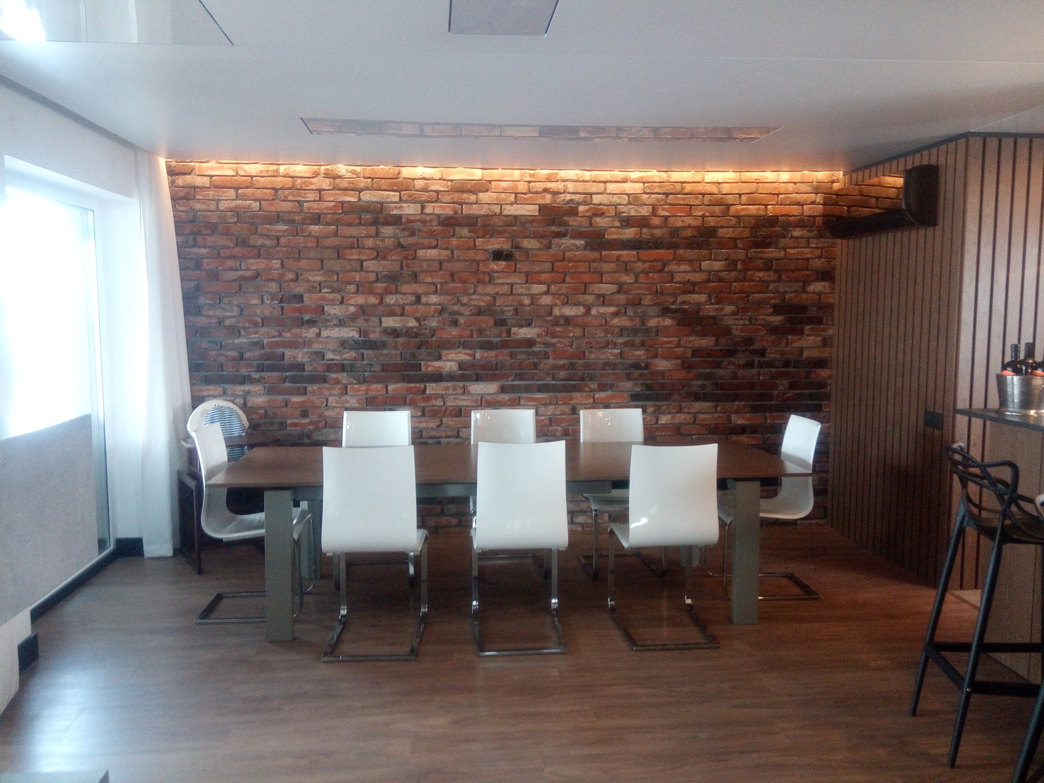 Jadalnia z ceglaną ścianą podświetloną od góry, stół z białymi krzesłami. Widoczne drewniane panele i bar z hokerami. Nowoczesne wnętrze po remoncie.