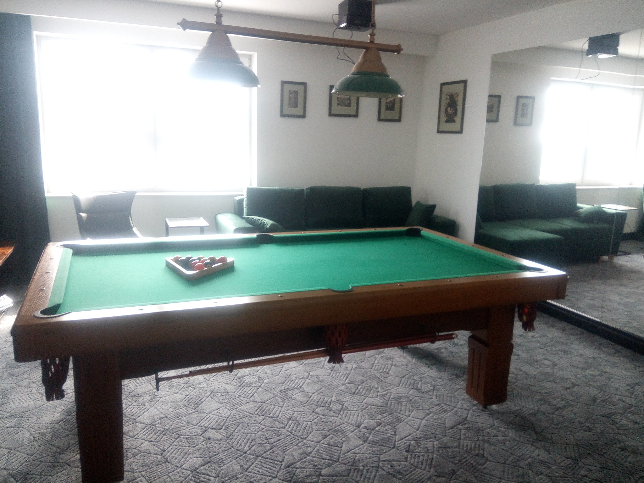 Stół bilardowy z zielonym suknem, ustawione bile, oświetlenie lampami. W tle kanapa i dekoracje ścienne. Widok na pokój z perspektywy gracza.