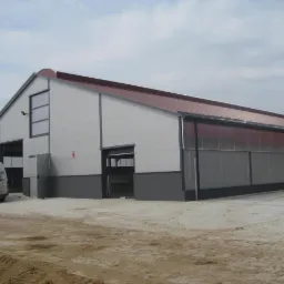 Gotowa hala z płyt warstwowych, z widocznym samochodem dostawczym firmy. Czerwony dach, szare ściany i brama wjazdowa. Hala na terenie budowy.