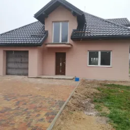 Niedawno wybudowany, jednopiętrowy dom z garażem w kolorze pudrowego różu z ciemnobrązowymi drzwiami i ciemnoszarą dachówką, otoczony brukowanym podjazdem i trawnikiem w trakcie zakładania...