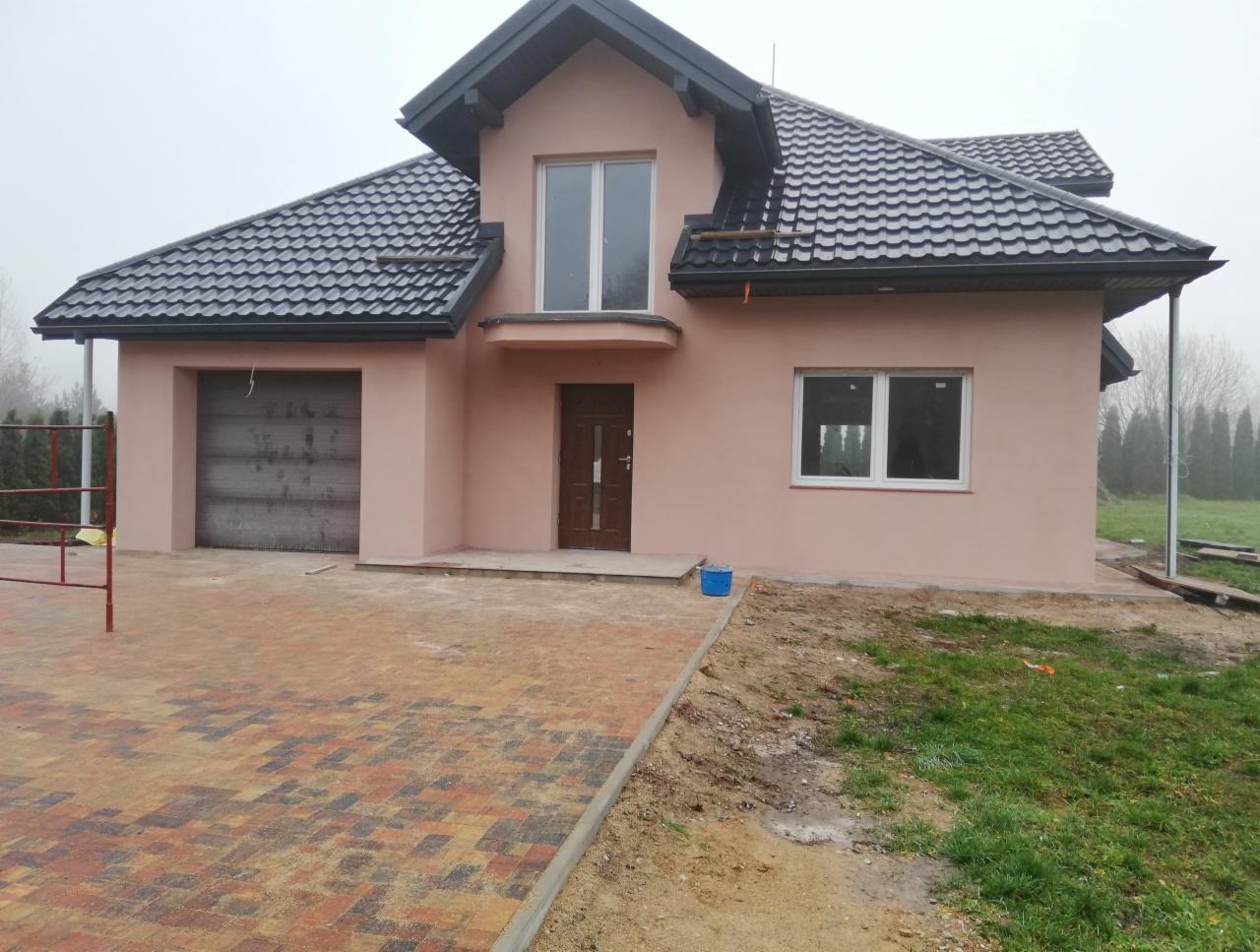 Niedawno wybudowany, jednopiętrowy dom z garażem w kolorze pudrowego różu z ciemnobrązowymi drzwiami i ciemnoszarą dachówką, otoczony brukowanym podjazdem i trawnikiem w trakcie zakładania...