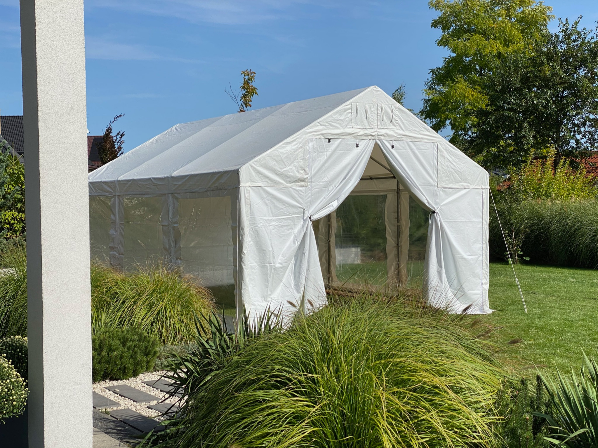 Biały namiot plenerowy z przezroczystymi ścianami bocznymi, ustawiony na trawniku w ogrodzie, otoczony ozdobnymi trawami i drzewami w słoneczny dzień.