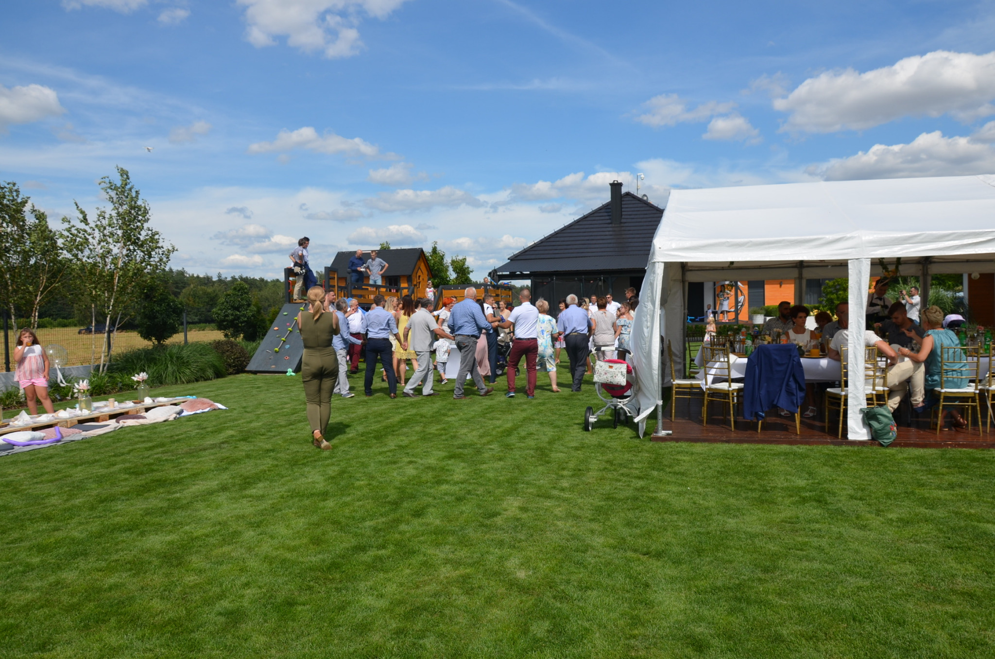 Plenerowa impreza z białym namiotem, gośćmi na trawniku, drewnianym domkiem zabaw z drabinką w tle i błękitnym niebem z chmurami.