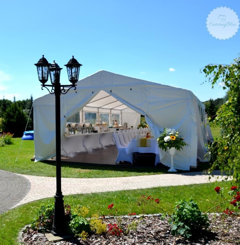 Biały namiot imprezowy z wyposażonym wnętrzem, widok z zewnątrz, oświetlony lampą uliczną, na trawniku z klombem kwiatowym.