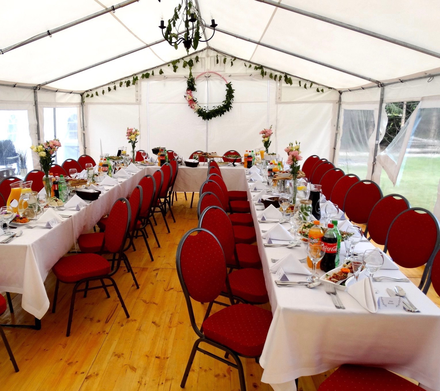 Uroczystość w białym namiocie: długie stoły z białymi obrusami, czerwone krzesła, dekoracje kwiatowe, napoje i zastawa. Podłoga z jasnego drewna. Dekoracyjny wieniec nad stołem.