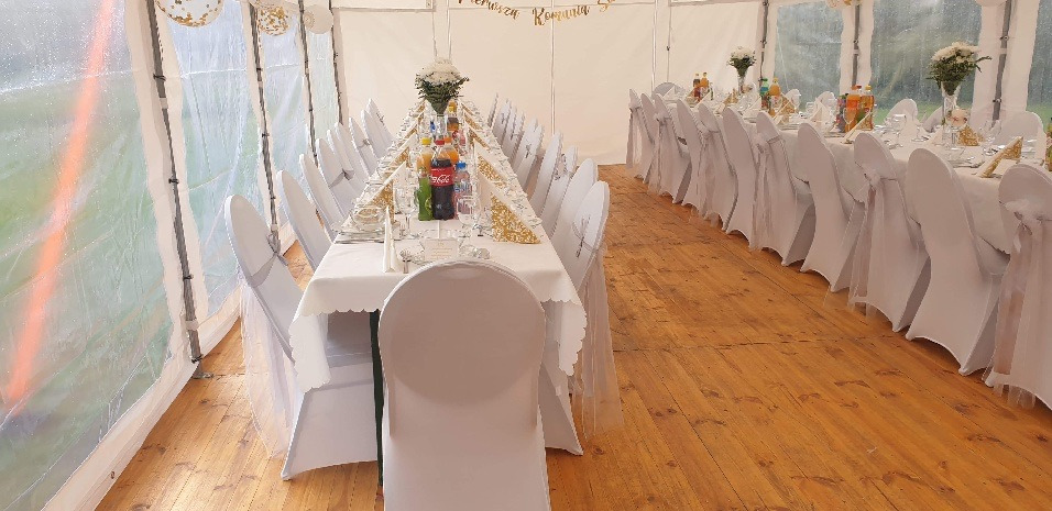 Wnętrze białego namiotu z drewnianą podłogą, przygotowane na przyjęcie komunijne. Długie stoły z białymi obrusami, nakryte złotymi serwetkami i dekoracjami kwiatowymi, otoczone krzesłami z białymi...