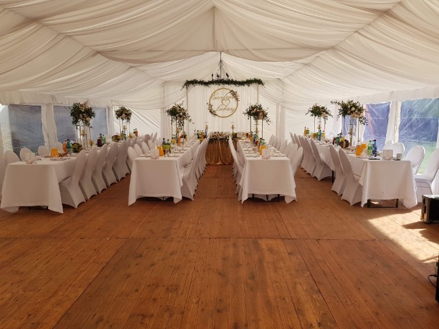 Uroczyste, długie stoły z białymi obrusami i krzesłami w białych pokrowcach, ustawione wewnątrz białego namiotu z dekoracjami kwiatowymi, przygotowane na przyjęcie w Łukowie. Drewniana podłoga...