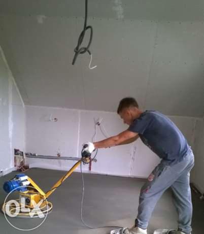 Pracownik w szarej koszulce i dresach wygładza wylewkę podłogową za pomocą elektrycznej zacieraczki w pomieszczeniu z surowymi ścianami.