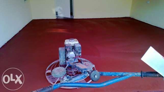 Czerwona wylewka podłogowa w trakcie wygładzania za pomocą zacieraczki mechanicznej, widoczna brama drzwiowa w tle.