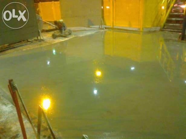 Świeżo wylana, mokra posadzka przemysłowa z widocznymi refleksami świetlnymi, maszyna do polerowania posadzek w tle.