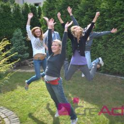 CreActive Team - Grupa młodych osób w dżinsach skacze na trawie przed żywopłotem, unosząc ręce w górę. Widoczne logo firmy w dolnej części kadru.