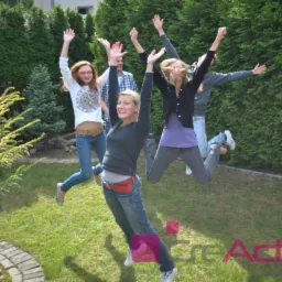 Grupa młodych osób w dżinsach skacze na trawie przed żywopłotem, unosząc ręce w górę. Widoczne logo firmy w dolnej części kadru.