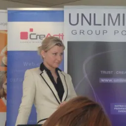 Kobieta w jasnym żakiecie na tle roll-upów reklamowych agencji interaktywnej. Widoczne logo 'CreActi' i 'Unlimited Group Poland'.