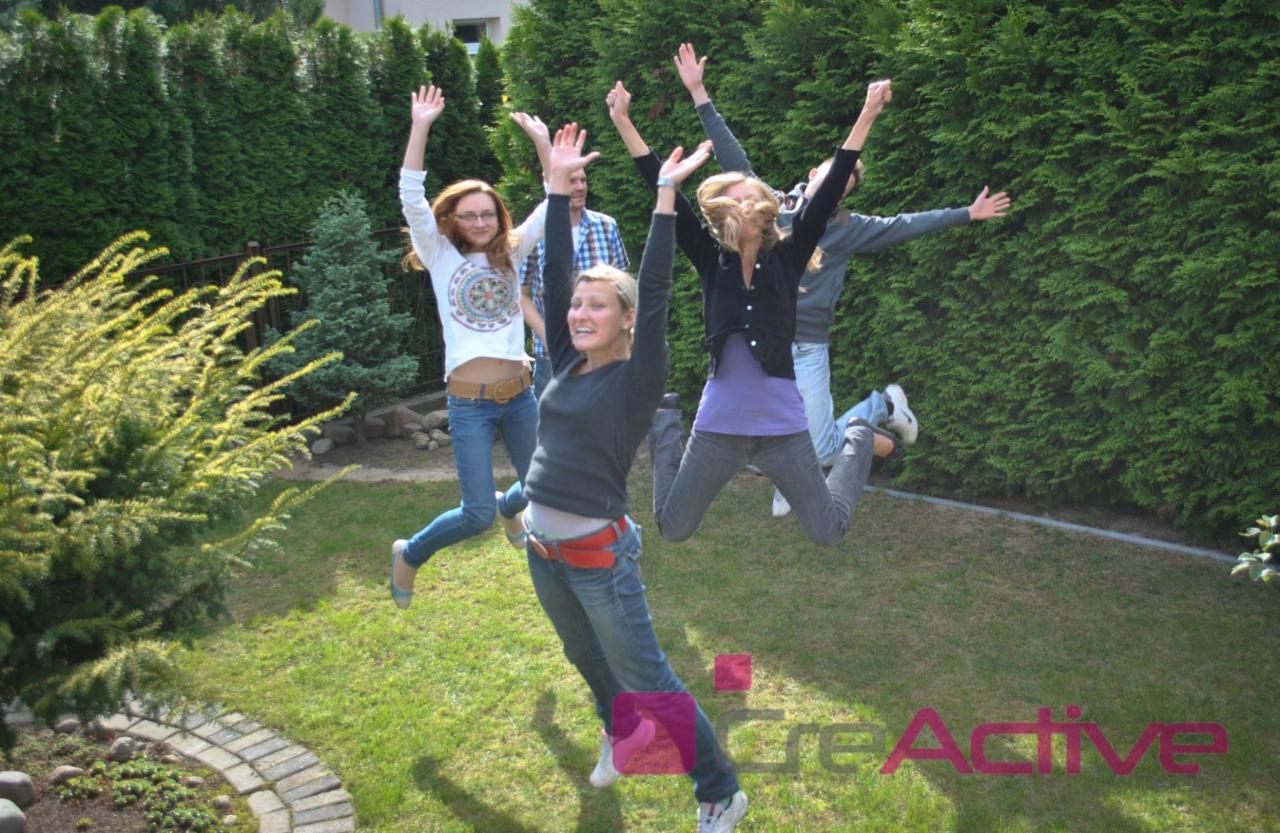 Grupa młodych osób w dżinsach skacze na trawie przed żywopłotem, unosząc ręce w górę. Widoczne logo firmy w dolnej części kadru.