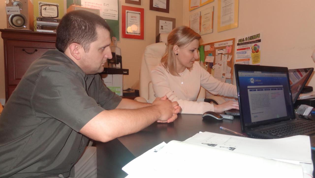 Dwie osoby pracują przy biurku z laptopami w biurze. Na ścianie wiszą ramki z certyfikatami i tablica korkowa. Jeden laptop wyświetla stronę internetową.