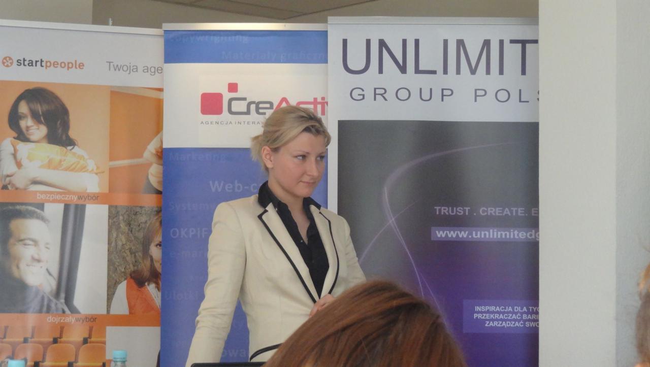 Kobieta w jasnym żakiecie na tle roll-upów reklamowych agencji interaktywnej. Widoczne logo 'CreActi' i 'Unlimited Group Poland'.