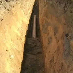 Wąski wykop w ziemi z ułożoną rurą kanalizacyjną z tworzywa sztucznego; widoczne ślady po pracy koparki, pryzma ziemi na górze wykopu i fragment budynku w tle.