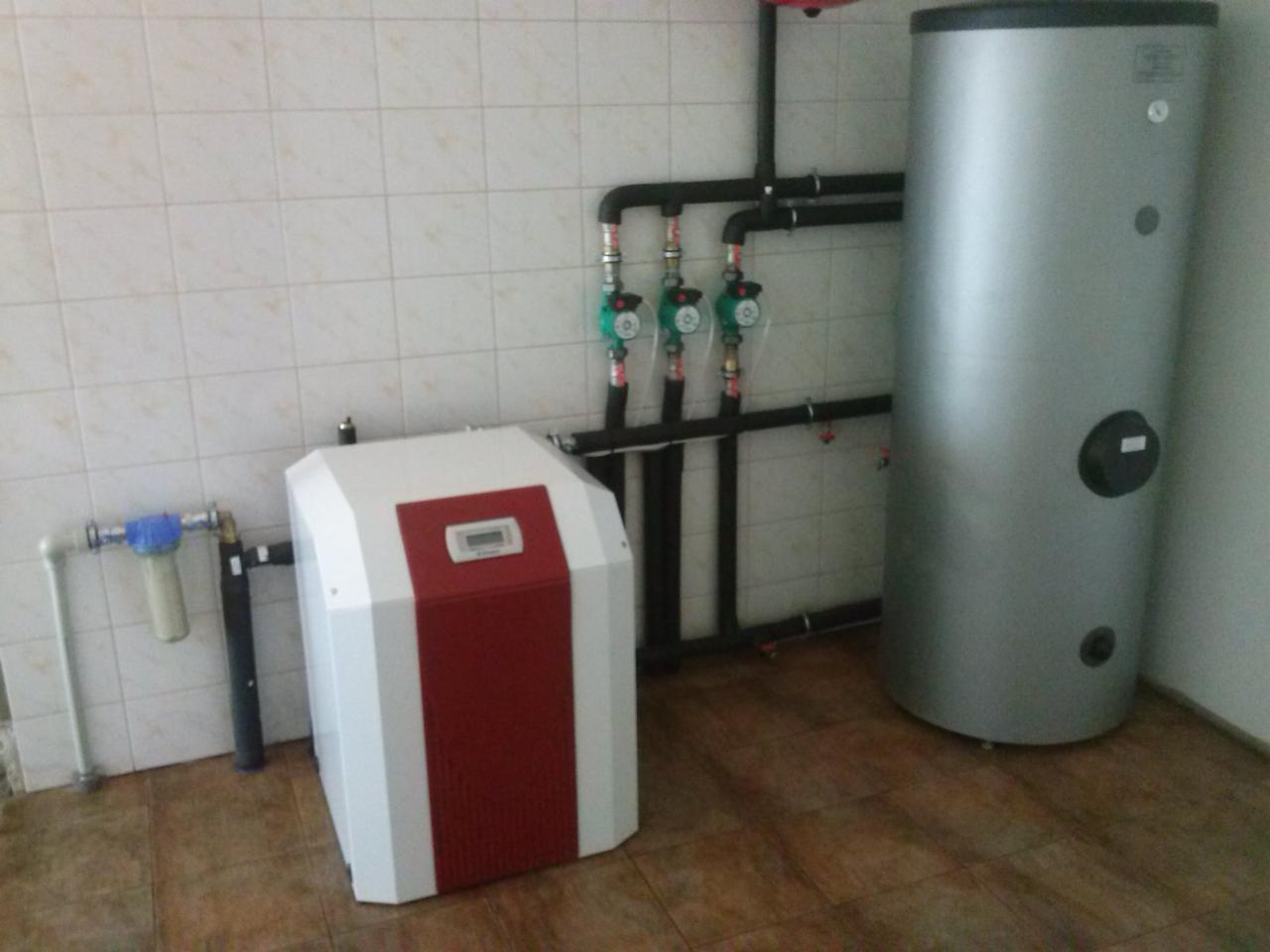 Biało-czerwona pompa ciepła z szarym zasobnikiem wody, rurami i filtrem na tle kafelkowanej ściany i brązowej podłogi. Widoczne zawory i elementy instalacji.