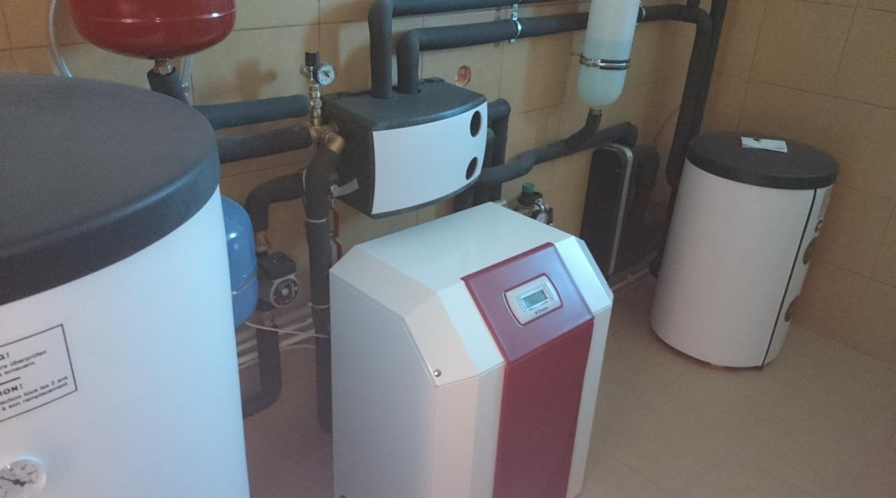 Zestaw urządzeń grzewczych w kotłowni: biały zasobnik ciepłej wody, pompa obiegowa, zbiornik wyrównawczy i rury instalacji grzewczej w pomieszczeniu z beżowymi płytkami.