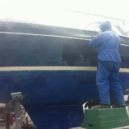 Pracownik w kombinezonie czyści kadłub jachtu strumieniem wody pod ciśnieniem, stojąc na zielonym podeście. Widoczne ślady czyszczenia i fragmenty nieoczyszczonej powierzchni.