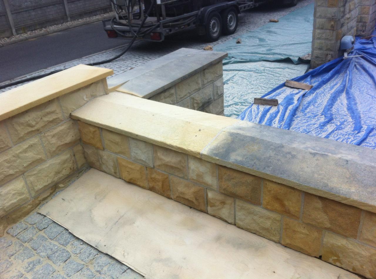 Fragment kamiennego murku, czyszczonego strumieniowo, z widoczną różnicą w kolorze między czystą a zabrudzoną powierzchnią. W tle przyczepa ze sprzętem.