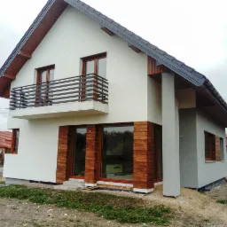 Dwukondygnacyjny dom jednorodzinny z białymi ścianami i drewnianą okładziną wokół okien, balkon z czarną balustradą, szara kolumna podtrzymująca dach, widok z zewnątrz na tle pochmurnego nieba...