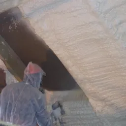 Pracownik w kombinezonie ochronnym natryskuje pianę izolacyjną na poddasze, widoczne krople piany w powietrzu, fragment rusztowania po lewej stronie.