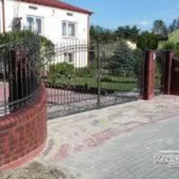 Oryginalne ogrodzenie z ceglanymi słupkami i metalową bramą, łukowe przejście, kostka brukowa, dom w tle. Nietypowe wykończenie.