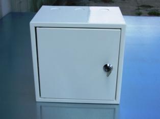 Biała, metalowa obudowa uniwersalna w kształcie sześcianu z drzwiczkami zamykanymi na klucz. Widoczne trzy okrągłe otwory na górnej ściance.