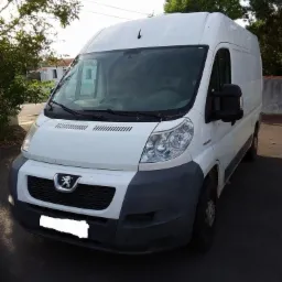 Biały bus Peugeot z szarym zderzakiem, zaparkowany na zewnątrz. Widoczny przód pojazdu z logo marki i reflektorami. Tło stanowią drzewa i fragment budynku.