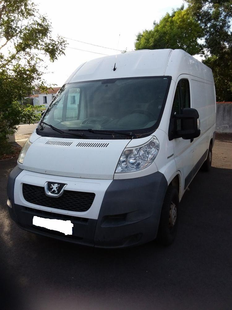 Biały bus Peugeot z szarym zderzakiem, zaparkowany na zewnątrz. Widoczny przód pojazdu z logo marki i reflektorami. Tło stanowią drzewa i fragment budynku.