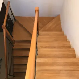 Widok z góry na drewniane schody z jasnymi stopniami i poręczą, z czarną metalową balustradą po jednej stronie, prowadzące na górę, z fragmentem drewnianej podłogi na górnym podeście ułożonej...