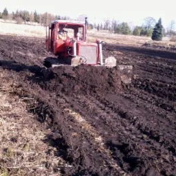 Czerwony traktor w trakcie niwelacji terenu w Szczecinku. Widoczna świeżo obrobiona ziemia i operator w kabinie, drzewa w tle.