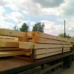 Świeżo pocięte, jasne belki drewniane, zabezpieczone pasami, ułożone na platformie transportowej samochodu ciężarowego, z widocznymi oznaczeniami wymiarów na końcach i kominem w tle.