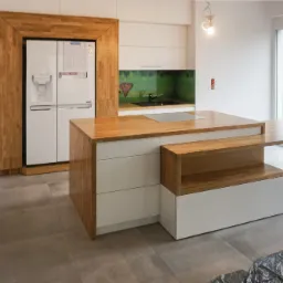 Nowoczesna kuchnia z wyspą i drewnianym blatem, białe fronty szafek, drewniana rama lodówki, widok na ogród przez duże okno, częściowo zakryta podłoga.