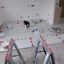 Układanie białych, wielkoformatowych płytek na podłodze. Widoczne poziomujące klipsy i kliny, torba narzędziowa, oraz plastikowe kozły malarskie.