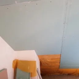 Montaż płyt gipsowo-kartonowych na suficie, widoczne łączenia i wkręty, fragment ściany pomalowanej na biało oraz zabezpieczona folią podłoga.