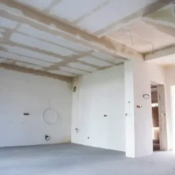 Surowe wnętrze w trakcie prac wykończeniowych: widoczne ślady po tynkowaniu na suficie, szare wylewki na podłodze, wystające kable i rury instalacyjne na ścianach, schody w stanie surowym.