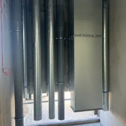 Wnętrze w trakcie instalacji systemu wentylacyjnego z widocznymi rurami spiralnymi, prostokątnym kanałem wentylacyjnym i rurami PCV, zamocowanymi do surowego betonowego stropu.