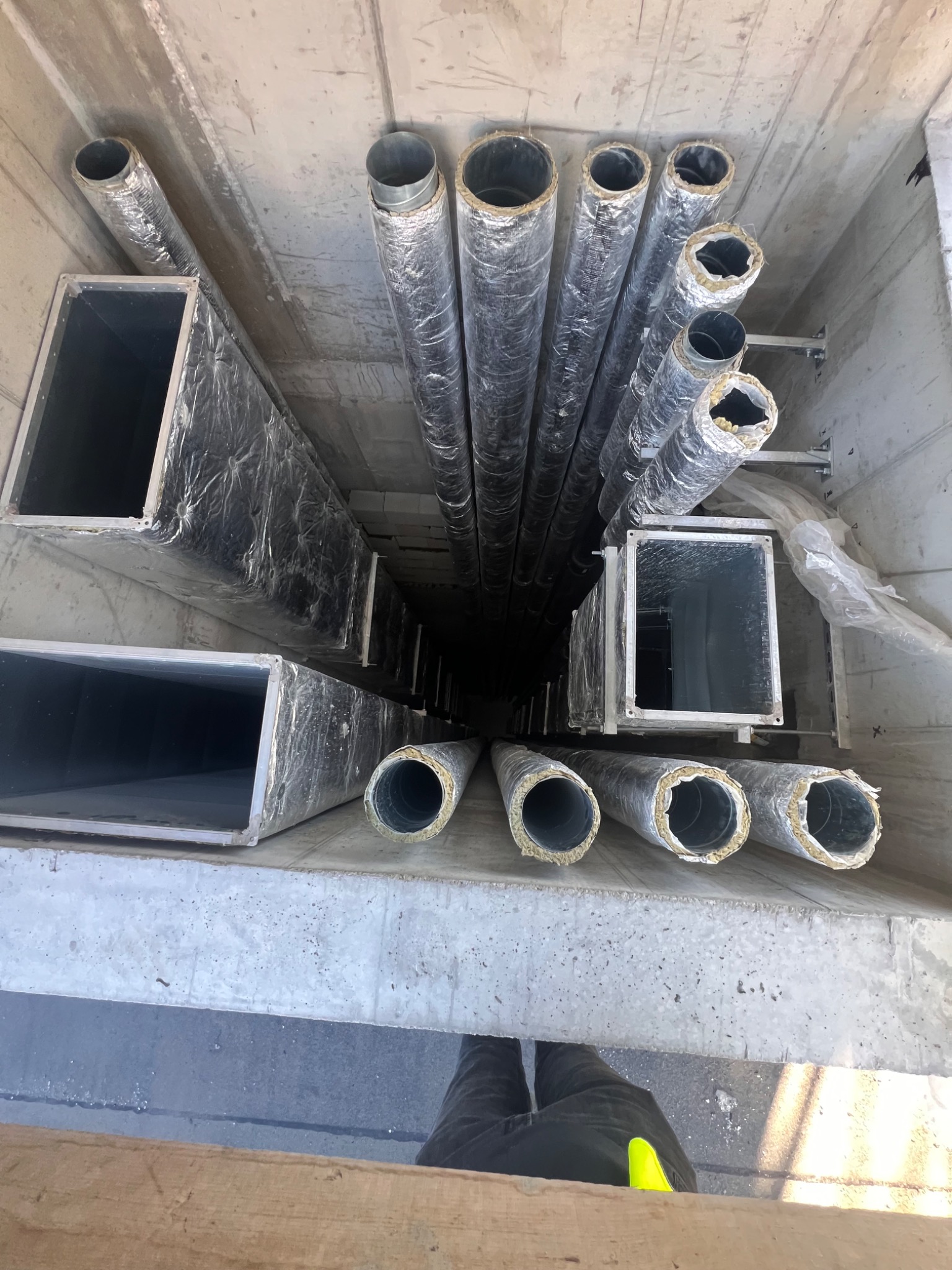 Wnętrze szybu z zamontowanymi, izolowanymi przewodami wentylacyjnymi o przekroju okrągłym i prostokątnym, widok z góry na dół, z fragmentem buta pracownika w rogu kadru.